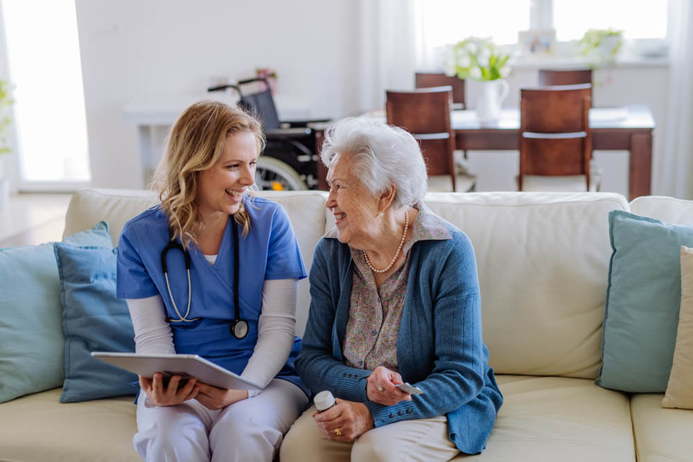 Female nurse sitting on a sofa with a memory care patient, laughing and conversing, at Traditions of Zanesville in Zanesville, OH.