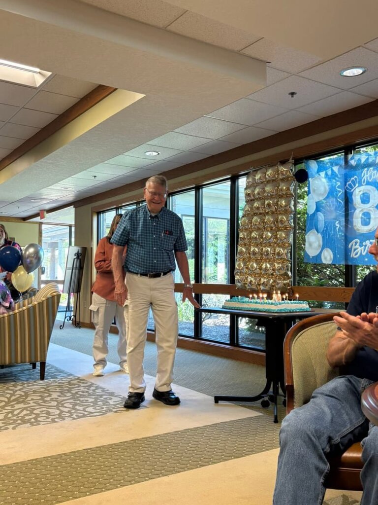 Community member Geoff smiles beside his lit birthday cake during his 80th birthday celebration at Traditions of Zanesville, with a blue "Happy 80th Birthday" banner and gold balloon display behind him.