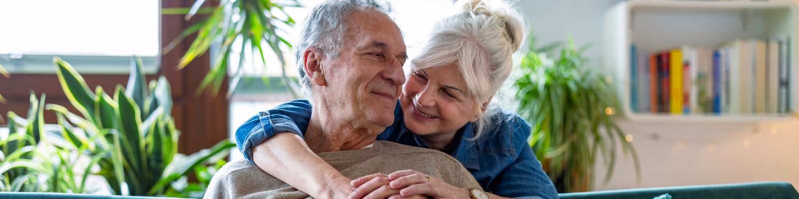 An older man sitting down, smiling at an older woman with white hair tied up wrapping her arms around his shoulders