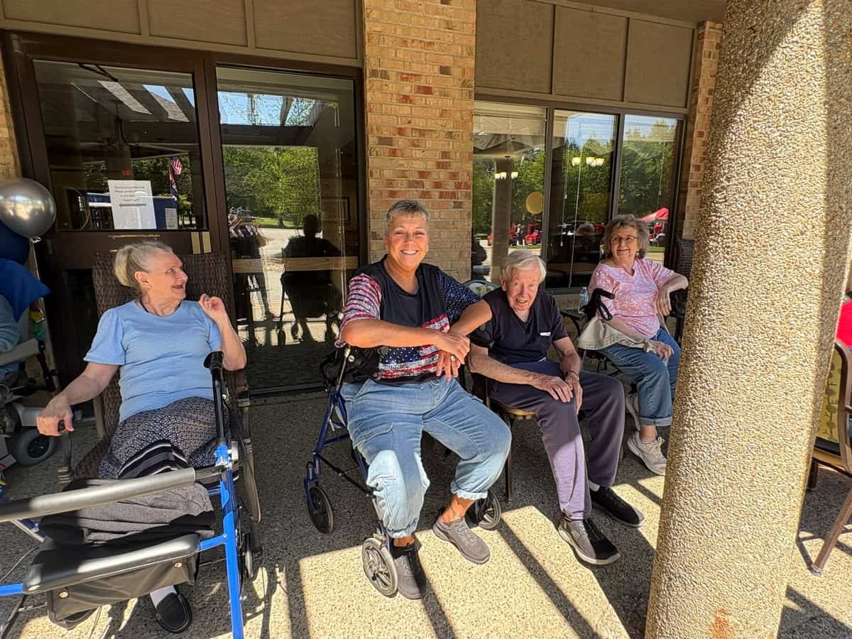 Three residents and a team member enjoy the view from the front entrance during the Traditions of Zanesville charity car show, with colorful show cars visible in the parking lot behind them.